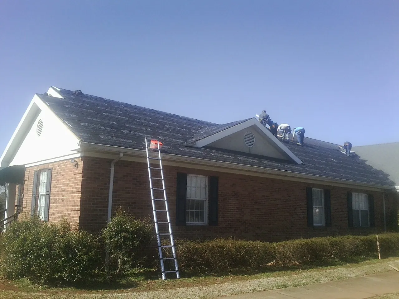 Crew tearing shingles off a brick ranch roof with a ladder set — Emergency Roof Repair work by our crew in Glenwood Landing