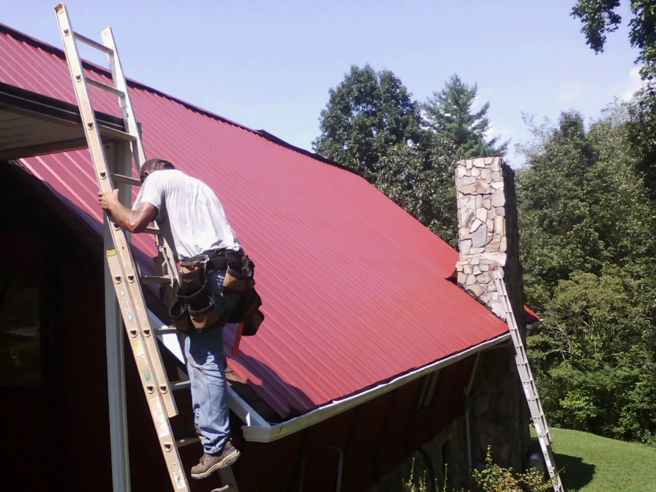 Roofer on a ladder beside a red metal roof and stone chimney — Soffit & Fascia Repair by Roofing Brothers in Glenwood Landing